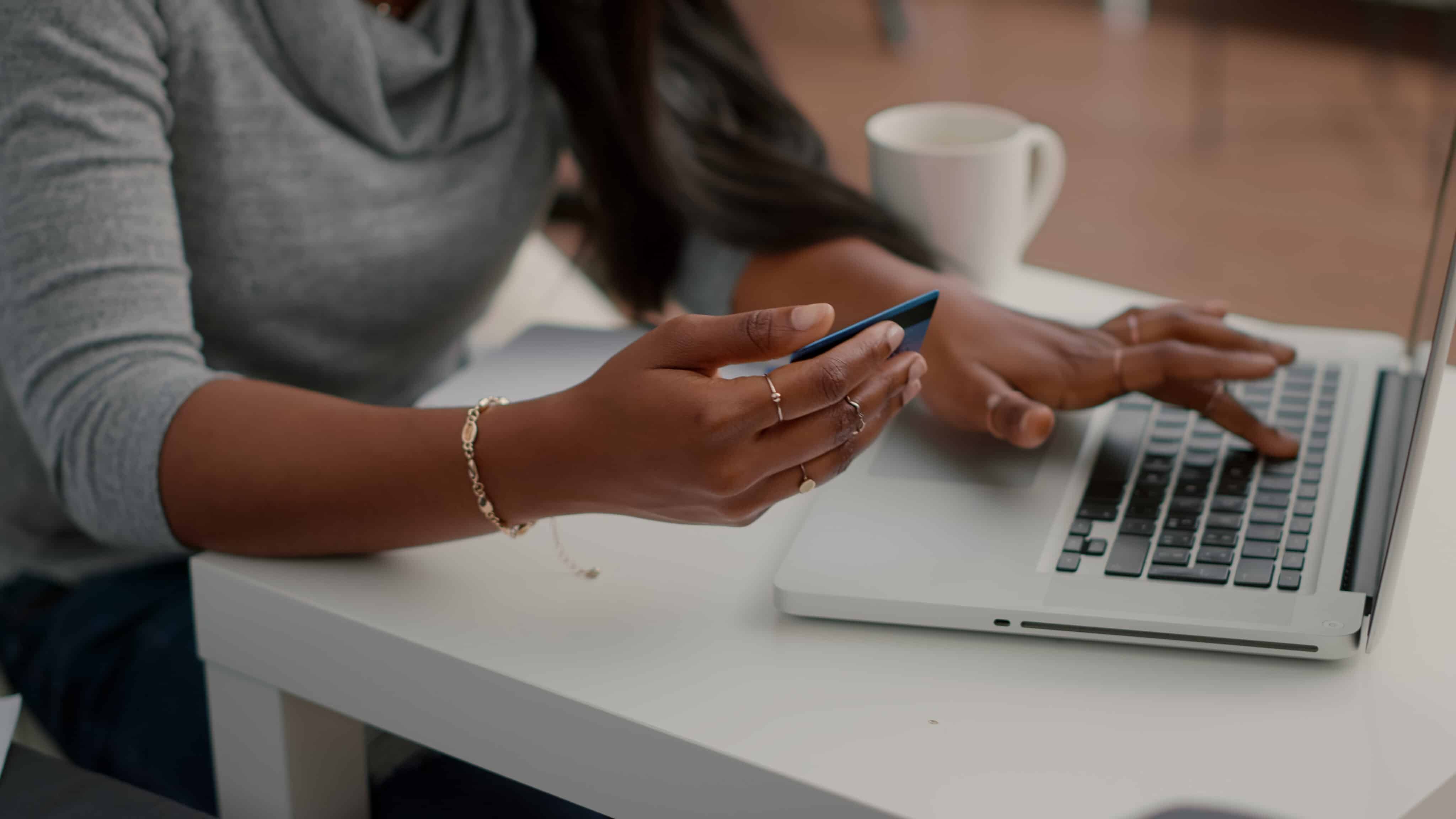 Black student holding cred card in hands doing electronic transcation searching online store, easy payment using digital gadget. Teenager browsing internet banking service on laptop in living room